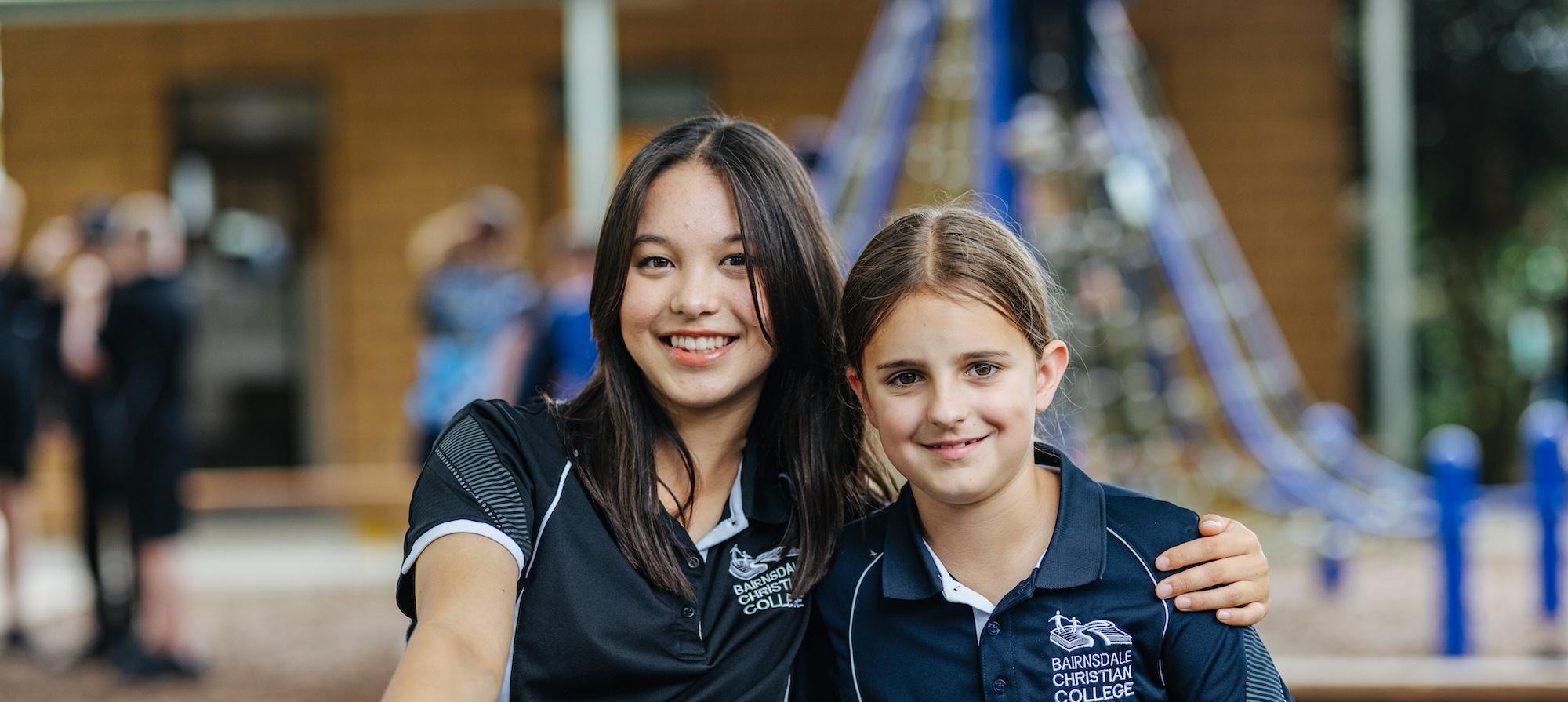 Two students smiling arm in arm in the school playground, representing friendship, belonging, and the warm welcome awaiting new families at Bairnsdale Christian College.